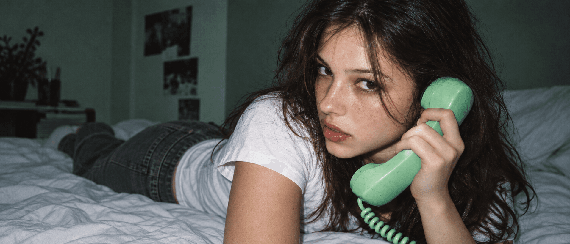 A young woman lying on a bed, holding a mint-green telephone handset to her ear and looking at the camera
